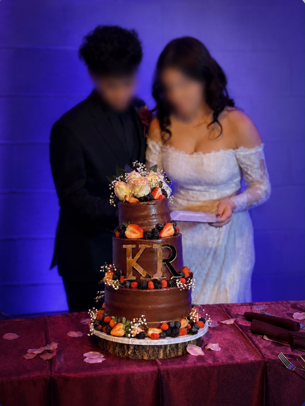 Three-tier chocolate ganache wedding cake with fresh berries, florals, and a gold KR monogram topper, with the newlyweds in the background